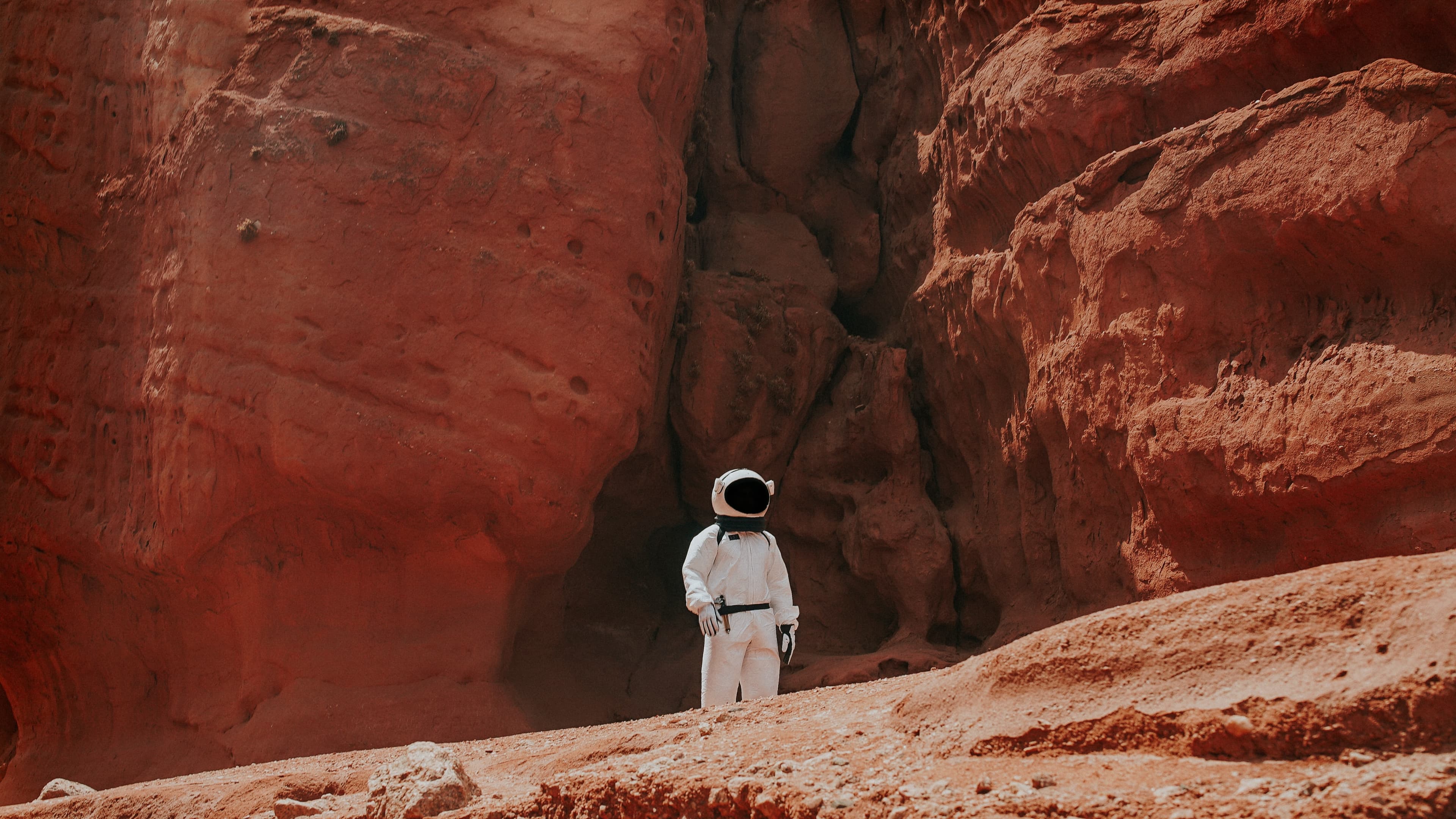 An astronaut in a white suit stands alone in a canyon with steep, red-rock walls.
